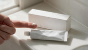 A plain white cream tube and a pea-size dot of ivermectin cream on a fingertip beside a 45 gram carton.